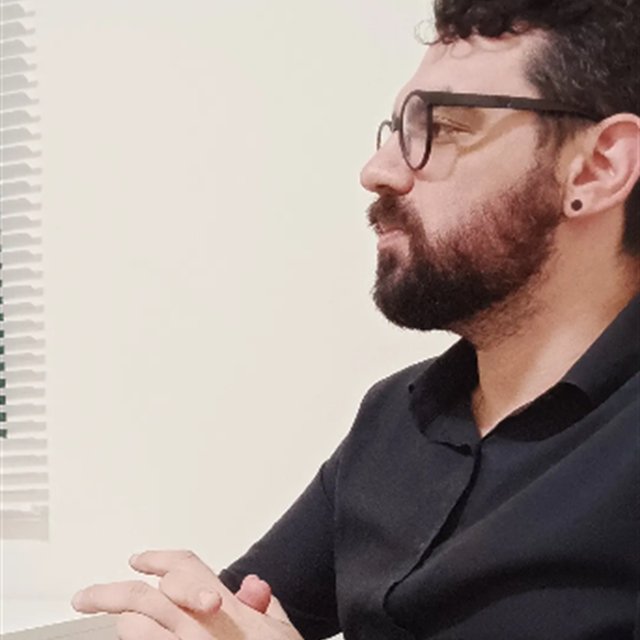 Felipe Queiroz em atendimento, de perfil, mãos entrelaçadas.